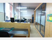 Moderner Büroarbeitsbereich mit Holzmöbeln, großen Fenstern und weiß gefliesten Wänden. Rechts befindet sich ein Konferenzraum mit Glaswänden, während der Hauptbereich Arbeitsplätze, Regale und Schreibtische umfasst.