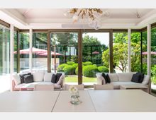 Moderner Innenraum mit großen Glasfenstern, die einen Blick auf einen gepflegten Garten mit runden Buchsbaumsträuchern und roten Sonnenschirmen bieten. Im Vordergrund stehen helle Sofas mit dunklen Kissen sowie ein Tisch mit einer Blumenvase.