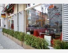 Außenansicht eines amerikanischen Diners mit großen Fenstern, hinter denen rote Sitzbänke und Tische sichtbar sind. Vor den Fenstern befinden sich eine Hecke in Holzpflanzkästen sowie Neon-Schilder mit Schriftzügen wie "OPEN".