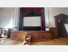 Bühnenraum in einem historischen Saal mit einer Holzverkleidung, einem kleinen Treppenpodest und roten Vorhängen, die eine weiße Leinwand einrahmen. Links und rechts stehen dekorative Pflanzentöpfe, die Decken und Wände sind mit weißen Stuckelementen verziert.