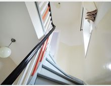 Treppe in einem hellen Innenraum mit abgerundeten Stufen, einem schwarzen Handlauf und einem Geländer mit weißen und orangefarbenen Streben. Im Hintergrund ist eine kleine Fensteröffnung mit hochgerolltem Vorhang sowie eine runde, hängende Deckenleuchte sichtbar.