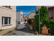 Schmale, gepflasterte Gasse in einem historischen Wohnviertel mit Häusern aus Putz und Klinkerfassaden; eine Hauswand ist dicht mit grünen Pflanzen bewachsen. Ein Straßenschild mit der Aufschrift „Pfarrgartenstr.“ ist sichtbar.