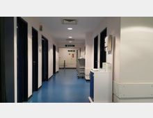 Korridor in einem Krankenhaus oder einer medizinischen Einrichtung mit blauem Boden, weißen Wänden und mehreren Türen auf beiden Seiten. Im Hintergrund befinden sich Notausgangsschilder, medizinisches Equipment und ein Wagen mit Utensilien auf der rechten Seite.