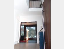 Moderner Innenraum eines Hauses mit offener Galerie und großen Fenstern, die den Blick auf Bäume freigeben. Im Vordergrund sind Fliesenboden, eine Holz- und Metallgeländerkonstruktion sowie eine blaue Kommode zu erkennen.