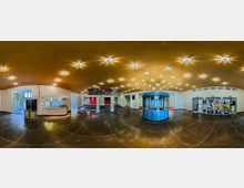 Panoramablick auf ein Foyer eines Theaters oder Kinos mit glänzendem schwarzen Boden. Zentral steht eine runde, blaue Kasse, umgeben von modernen Leuchten an der goldfarbenen Decke, mit roten Vorhängen im Hintergrund und Bildern an Glastüren auf der rechten Seite.