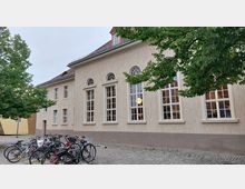 Außenansicht eines hellen Gebäudes mit hohen, rechteckigen Fenstern und kleinen, halbkreisförmigen Oberlichtern, flankiert von einem Baum auf der linken Seite. Im Vordergrund stehen zahlreiche abgestellte Fahrräder auf einem gepflasterten Platz.