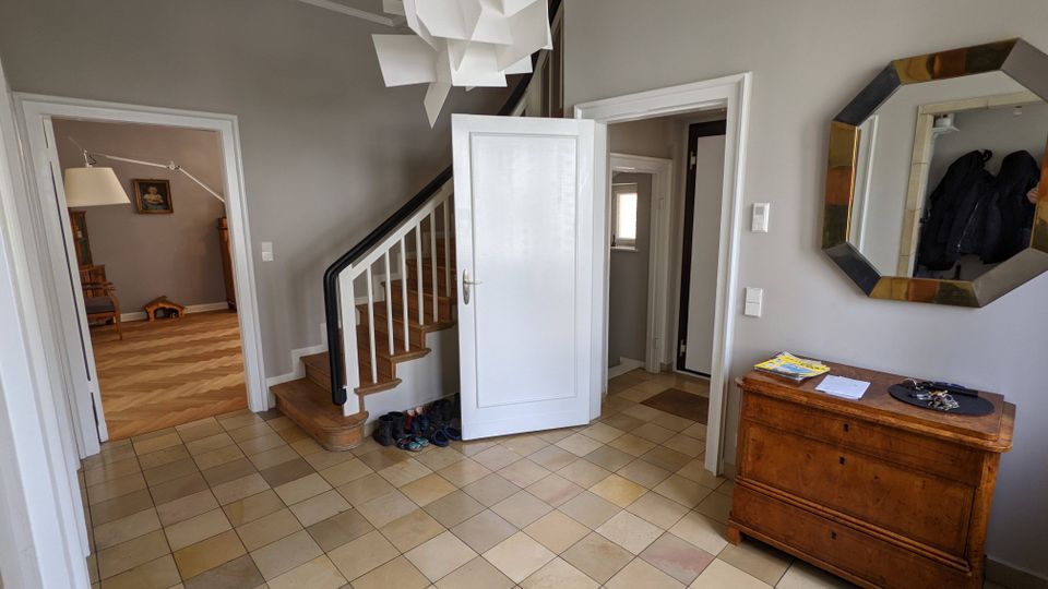 Heller Eingangsflur eines Wohnhauses mit großformatigen beigefarbenen Bodenfliesen, grauen Wänden und einer Holztreppe mit weißem Geländer. Mehrere offene Türen führen in angrenzende Räume; an der Wand hängen ein achteckiger Spiegel, daneben steht eine Holzkommode, und an der Decke hängt eine skulpturale weiße Pendelleuchte.