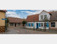 Eine gepflasterte Dorfstraße mit kleinen Wohnhäusern, darunter ein weißes Haus mit blauen Fensterläden und einer blauen Tür. Im Vordergrund stehen einzelne Bäume, eine Bank und eine Straßenlaterne, daneben ein Straßenschild mit der Aufschrift „Klosterstraße“.