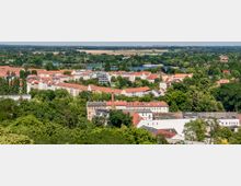 Luftaufnahme einer Stadt mit roten Ziegeldächern, umgeben von dichtem Grün und Bäumen. Im Hintergrund sind ein großes Gewässer sowie weite Felder und Wälder sichtbar.