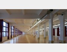 Großer, heller Innenraum mit Holzboden, weißen Säulen und langen Reihen von Leuchtstofflampen an der Decke. Auf der linken Seite befinden sich große Fenster mit roten Vorhängen, die Tageslicht hereinlassen.