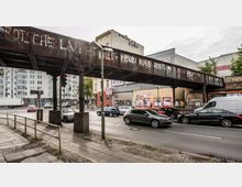 Straßenszene in einer urbanen Umgebung mit einer erhöhten, rostigen Eisenbahnbrücke, die von Pfeilern getragen wird und Graffiti trägt. Darunter verlaufen mehrspurige Fahrbahnen mit Autos, umgeben von Gebäuden, darunter ein Supermarkt mit Werbeschildern.
