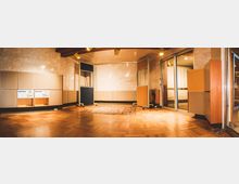 Ein Tonstudio mit hölzernem Parkettboden, akustischen Wandpaneelen und einer Trennwand aus Glas und Holzrahmen. Die Wände sind mit warmen, beigen Materialien verkleidet, und die Decke ist mit hölzernen Lamellen ausgestattet, unter denen Beleuchtung montiert ist.