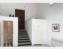 Innenraum mit weiß gestrichenen Wänden, einer offenen Treppe aus grauem Stein, die zu einer braunen Holztür führt. Links befindet sich ein offenes Badezimmer mit Fliesenboden, rechts eine weiße Tür mit Toilettensymbol und ein Heizkörper unter einem gerahmten Bild.