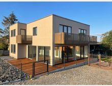 Modernes zweistöckiges Wohnhaus mit flacher Dachkonstruktion, großen Glasfenstern und Balkonen mit Metallgeländern. Das Haus steht auf einem gepflasterten Grundstück mit einem kleinen Zaun im Vordergrund.