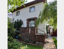 Zweistöckiges Wohnhaus mit heller Fassadenfarbe und roten Klinkerelementen, umgeben von einem gepflegten Garten mit Rasen, Sträuchern und Bäumen. Ein gepflasterter Weg führt entlang des Hauses, und ein weißer Fensterbereich ist von Klinkersteinen eingerahmt.