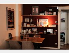 Gemütlicher Essbereich in einer Wohnung mit Holztisch und Polsterstühlen vor einer dunkelroten Wand mit drei langen Wandregalen voller Bücher, Bilderrahmen, Vasen und einer kleinen Lampe. Links hängt ein gerahmtes Poster, rechts führt eine Türöffnung in ein angrenzendes Zimmer.