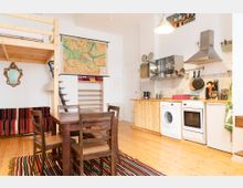 Wohnbereich mit offener Küche in einem hellen Raum. Zu sehen sind ein Holztisch mit Stühlen, ein Hochbett aus Holz, ein Wandspiegel, Regale, eine kleine Küche mit Holzelementen sowie Herd, Waschmaschine und Kühlschrank; eine Karte von Brandenburg hängt an der Wand.