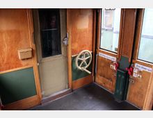 Holzvertäfeltes Interieur eines historischen Zugabteils mit grünem Türbereich, runden Fenstern und einem großen Drehgriff an der Tür. Schilder mit der Aufschrift „Nicht öffnen, bevor der Zug hält!“ sind an den Türen angebracht.