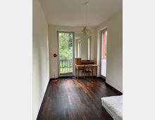 Ein schlichtes Zimmer mit dunklem Holzboden, weißen Wänden und einer Glas-Balkontür, die einen Blick ins Grüne bietet. Im Raum stehen ein Holzschreibtisch mit Stuhl, eine rote Stehlampe und ein Bett am rechten Bildrand.