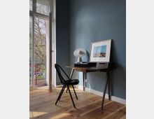 Ein kleiner, minimalistischer Arbeitsbereich in einem Raum mit blaugrauer Wand, einem schmalen Holztisch, schwarzem Stuhl und Tischlampe. Links führt eine geöffnete Balkontür mit Blick auf eine mit Bäumen gesäumte Außenanlage.