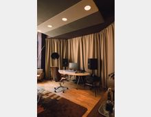 Arbeitszimmer mit einem weißen Schreibtisch, auf dem ein Monitor, Studio-Lautsprecher und ein MIDI-Keyboard stehen. Im Hintergrund beige Vorhänge, eine abgehängte Decke mit integrierter Beleuchtung und ein Holzboden mit einem großen Teppich.