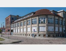 Gebäude im Stadtbereich mit hellbeiger Fassade, großen Fenstern und teilweise abgedeckten Scheiben. Es steht an einer gepflasterten Straße, im Hintergrund ist ein roter Kirchturm sichtbar.