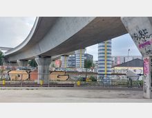 Eine graue Autobahnbrücke mit breiten, gebogenen Betonträgern überspannt eine städtische Umgebung. Die Brückenpfeiler sind in blau-weißen Streifen gestaltet, im Hintergrund sind Gebäude, Werbeschilder und Parkplätze sichtbar. Im Vordergrund stehen ein Geländer und ein mit Graffiti bemalter Betonsockel.