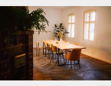 Heller Speiseraum mit einem langen Holztisch, umgeben von Holzstühlen und einem gepolsterten Sessel. Der Raum hat hölzernes Fischgrätenparkett und große Fenster, durch die Tageslicht einfällt, sowie dekorative Pflanzen im Hintergrund.