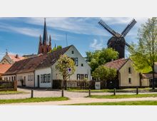 Das Bild zeigt eine ländliche Szene mit traditionellen Wohnhäusern mit roten Ziegeldächern im Vordergrund. Im Hintergrund stehen eine historische Holzwindmühle sowie ein spitzer Kirchturm, umgeben von Bäumen unter einem blauen Himmel.