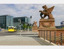 Straßenszene in einer Stadt mit Blick auf ein modernes Gebäude mit Glasfassade, im Hintergrund als Hauptbahnhof erkennbar. Im Vordergrund befinden sich zwei dekorative Sandsteinfiguren in Form geflügelter Kreaturen auf einem Geländer sowie eine gelbe Stadtbuslinie an einer Kreuzung mit Ampeln.