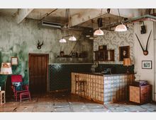 Innenraum einer rustikalen Bar mit industriellem Stil, einer Theke aus gemusterten Fliesen, grünen Kacheln an den Wänden und einer Mischung aus antiken Möbeln und Dekorationen wie Tierköpfen, Masken sowie Porträts. Hängelampen und freiliegende Kabel verlaufen unter einer abgenutzten Decke, der Holzfußboden ist mit bunten Akzenten gestaltet.