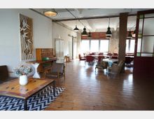 Gemütlicher Innenraum mit Holzboden und einer Mischung aus Vintage- und modernen Möbeln, darunter lederne und geflochtene Sessel, eine Holzanrichte, ein dekorativer Couchtisch und rote Stühle an runden Tischen im Hintergrund. Der Raum wird von großen Fenstern mit Holzjalousien und mehreren Pendelleuchten beleuchtet.