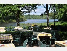 Außenterrasse eines Cafés oder Restaurants mit gelb-weiß karierten Tischdecken auf grünen Kunststoffstühlen, umgeben von Bäumen und Hecken, mit Blick auf einen See und eine bewaldete Landschaft im Hintergrund. Eine Laterne steht zwischen den Sitzbereichen.