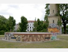 Rundes, niedriges Backsteingebäude mit Graffiti an den Wänden, umgeben von Bäumen und einer Wiese; im Hintergrund ein historischer, rechteckiger Turm aus gelbem Backstein, ebenfalls teilweise mit Graffiti. Im Hintergrund sind Wohnhäuser mit hellen Fassaden sichtbar.