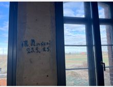 Eine detailreiche, rustikale Mauer mit verblasstem Schriftzug ("23.5.25") steht neben einem großen Fenster mit schwarzem Metallrahmen, das den Blick auf eine weite Landschaft mit Feldern, einem roten Gebäude und einem bewölkten Himmel bietet.