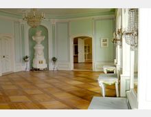 Heller, historischer Raum mit hellgrünen Wänden, verziert mit weißen Stuckelementen. Ein zentral platzierter, dekorativer Kachelofen, ein edler Kronleuchter, Parkettboden und durchgehende Fensterspiegel prägen den Raum; im Hintergrund ist eine geöffnete Tür zu einem angrenzenden Raum sichtbar.