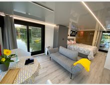 Modernes, helles Zimmer mit großen Glas-Schiebetüren, die den Blick auf einen See bieten. Der Raum verfügt über eine graue Couch, ein Doppelbett, einen kleinen Esstisch mit Tischdecke und Blumendekoration und einen Boden in Holzoptik.