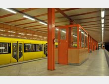 U-Bahnhof mit gelbem Zug an einem Bahnsteig, umgeben von orangen Stahlträgern und Wänden. Im Vordergrund ein orangenes Wachhäuschen, im Hintergrund verlaufen helle Deckenleuchten entlang des Bahnsteigs.