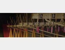 Ein alter Garderobenbereich mit nummerierten Hakenleisten aus Metall, an denen gelbe Gurte und Schlüsselanhänger in Rosa und Blau hängen. Die Beleuchtung ist gedämpft, und der Hintergrund ist dunkel.