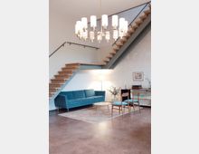 Ein modernes Wohnzimmer mit minimalistischem Design, bestehend aus einer blauen Couch, einem kleinen runden Beistelltisch und zwei Holzstühlen mit blauen Polstern. Über dem Raum hängt ein auffälliger Kronleuchter, und eine Treppe mit Holzstufen verläuft an zwei Wänden entlang.