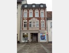 Fassadenansicht eines dreistöckigen Gebäudes in historischem Stil mit rotem Backstein und weißen Stuckverzierungen, darunter ein Detail mit der Aufschrift „Erbaut 1907“. Im Erdgeschoss befinden sich Geschäfte, darunter ein Friseursalon und ein Telekom-Laden, und vor dem Gebäude steht ein Fahrrad.