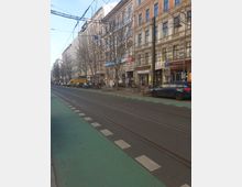 Straßenszene mit Wohn- und Geschäftshäusern in historischer Architektur, einer zweispurigen Straße mit Straßenbahnschienen sowie einem grünen Fahrradweg. Vor den Gebäuden stehen Bäume, parkende Autos und Fahrräder, während Geschäfte, Cafés und Schilder die Ladenfronten säumen.