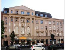 Historisches Gebäude mit klassizistischer Fassade, gekennzeichnet durch hohe Säulen, große Rundbogenfenster und ein Dreiecksgiebel. Im Erdgeschoss befinden sich mehrere Geschäfte und Restaurants, darunter ein Lokal mit italienischer Beschriftung, während davor parkende Autos und Bäume stehen.