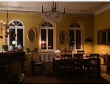 Ein stimmungsvoll beleuchtetes Wohnzimmer mit gelben Wänden, drei großen Bogenfenstern, einem Kronleuchter und einem Esstisch mit Holzstühlen. Dekorationen wie Kerzen, Weihnachtsschmuck und Spiegel prägen den Raum, der an eine gemütliche Weihnachtszeit erinnert.