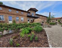 Backsteingebäude mit zwei Etagen und weißen Sprossenfenstern, umgeben von einem Garten mit Rosenbüschen, Steinwegen und Holzmulchbeeten. Im Hintergrund ein weiteres Gebäude und ein Schornstein, die zu einem Hofensemble gehören.