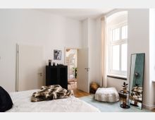 Schlafzimmer mit weiß gestrichenen Wänden, einem großen Fenster mit Vorhang und Holzboden. Im Vordergrund ein Bett mit Fellimitatdecke, ein schwarzes Regal an der Wand, ein langer Spiegel am Boden und eine Tür, die in ein weiteres Zimmer führt.