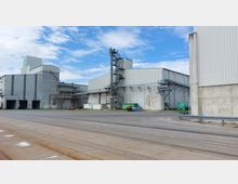 Industrieanlage mit großen grauen Lagerhallen aus Metall und Beton, umgeben von offenen Flächen. Im Vordergrund sind Schienen und asphaltierte Wege zu sehen, während sich im Hintergrund Förderanlagen, Container und ein grüner Tank befinden.