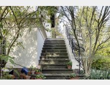 Außentreppe eines hellen Gebäudes mit schmiedeeisernem Geländer, umgeben von Bäumen und Pflanzen; am unteren Treppenabsatz stehen mehrere Blumentöpfe.