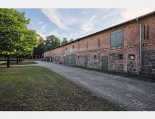 Roter Backsteinbau mit grauen Steintoren und -fenstern, flankiert von einem Pflasterweg und einer Grünfläche mit Bäumen auf der linken Seite, im Hintergrund umgeben von dichtem Baumbestand unter einem wolkigen Himmel.