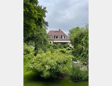 Ein Einfamilienhaus mit rotem Ziegeldach, umgeben von einem üppigen Garten mit Bambus, Blumen und hohen Gräsern. Im Vordergrund ist eine Rasenfläche mit Gartenstühlen sichtbar.